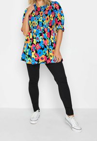 Nainen, jolla on päällään värikäs kukallinen paita puffihelmilla, mustat istuvat housut ja valkoiset lenkkarit, seisoo käsi kasvojaan lähellä.