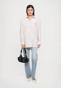 Mulher a usar uma camisa oversized às riscas rosa e branca, jeans claros com manchas rosas, sapatos de salto brancos, segurando uma mala preta com studs.
