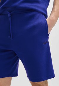 Paarse shorts van een gestructureerde stof met een elastische tailleband en trekkoord. Bevat zijzakken en een klein logo op het been.