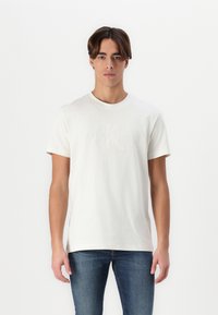 Bílá bavlněná tričko s vystouplým logem Calvin Klein na přední straně, kulatým výstřihem a krátkými rukávy. Nosené s modrými džíny.
