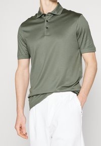 Olivgrön polo skjorta i slätt tyg med krage, tre svarta knappar och korta ärmar. Matcher med vita byxor.