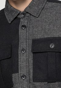 Sort og grå skjorte med knaplukning og ternet mønster, der har en uld-agtig tekstur og to forreste lommer med klaplukning.