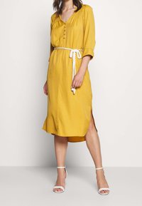 Musztardowa sukienka z rękawami 3/4, z detalami na guzikach, bocznymi rozcięciami i białym sznurkiem w talii; gładka faktura materiału. Białe szpilki.