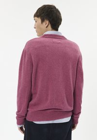 Rózsaszín kötött pulóver bordázott gallérral és mandzsettával, laza szabással és finom textúrával, kék-fehér csíkos ing fölé rétegezve.