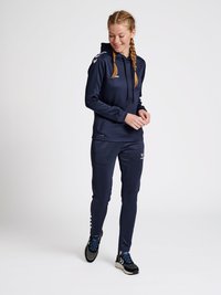 Navy atletiek tracksuit met een capuchon. Heeft een gladde textuur, elastisch materiaal en contrasterende witte schouderaccenten. Sneakers met blauwe details.
