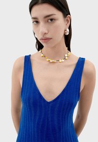 Robe sans manches rayée bleue avec un décolleté en V profond. Le mannequin porte un collier à perles jaunes et blanches et des boucles d'oreilles en argent.