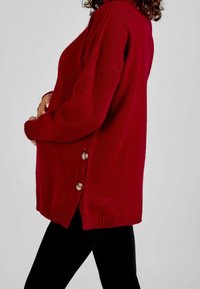 Suéter de punto burdeos con un ajuste holgado, presenta botones laterales y puños acanalados. Textura suave y dobladillo que cae por debajo de las caderas.