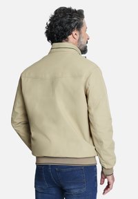 Chaqueta bomber beige con cuello alzado, puños y cinturilla de canalé. Textura suave, detalles mínimos y costura en el yugo trasero.