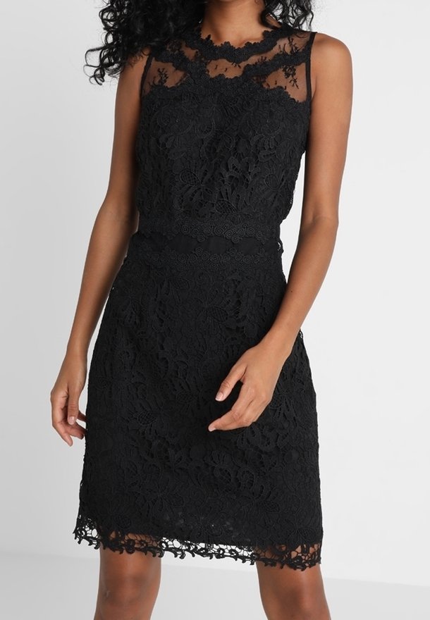Femme portant une robe noire en dentelle sans manches avec un décolleté transparent et des motifs floraux, se tenant devant un fond blanc uni.