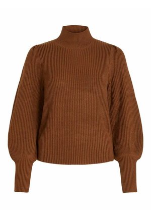Kvinde iført brun ribstrikket sweater med puffede ærmer og sorte højtaljede bukser foran en ensfarvet baggrund.