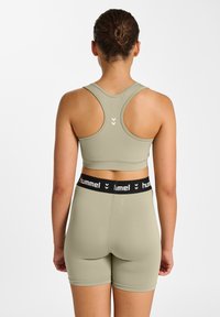 Oliwkowy zielony sportowy biustonosz i pasujące krótkie spodenki. Biustonosz ma design z wycięciem na plecach typu racerback; spodenki posiadają czarną taśmę z białym logo. Gładka tekstura.