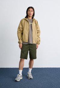 Lys brun jakke, grå T-skjorte, grønne shorts, hvite sokker og flerfargede joggesko. Bakgrunnen er enkel og teppet er blått.