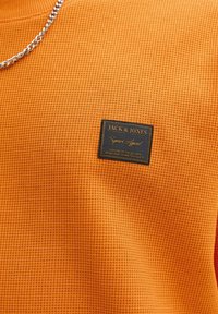 Oranje textiel met een kleine rechthoekige donkere label met het merk "Jack & Jones" en gouden tekst.