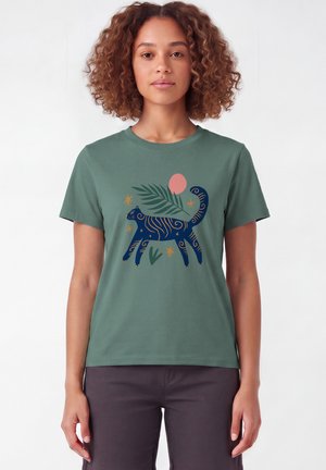 Femme aux cheveux bouclés portant un t-shirt vert avec un chat bleu à rayures orange, des feuilles vertes, un cercle rose et des étoiles jaunes.