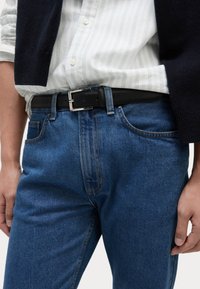 Cinturón de cuero negro con una hebilla plateada llevado sobre unos vaqueros de mezclilla azul. Camisa a rayas blancas visible en la parte superior, mostrando un abrigo de punto en capas.