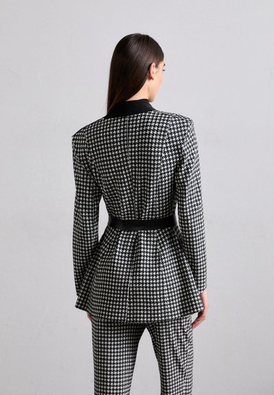 Fekete-fehér houndstooth mintás blézer peplum sziluettel, hosszú ujjakkal és fekete gallérral, amelyet derékon öv szorít.