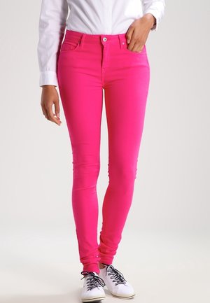 Personne portant un jean slim rose vif, une chemise blanche boutonnée et des baskets blanches avec des lacets noirs, debout devant un fond uni.