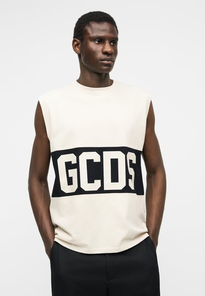 Άνδρας με κοντά κατσαρά μαλλιά φοράει αμάνικο λευκό πουκάμισο με έντονο μαύρο λογότυπο "GCDS" στο στήθος, τα χέρια στις τσέπες, μπροστά σε απλό φόντο.