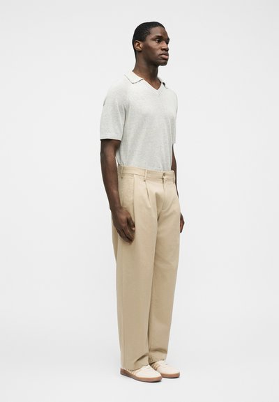 Άνδρας που στέκεται με μπεζ πλισέ παντελόνι, γκρι ανοιχτό κοντομάνικη πλεκτή μπλούζα και κρεμ αθλητικά παπούτσια μπροστά σε απλό λευκό φόντο.