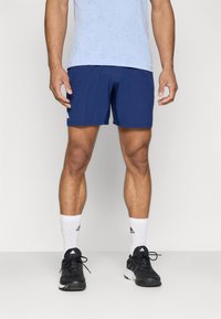 Shorts de sport bleus, en tissu léger, longueur mi-cuisse, avec un motif texturé. Baskets noires avec des accents blancs, chaussettes blanches à mi-mollet.
