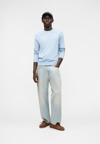 Pull bleu clair, jean bleu clair ample et chaussures bateau marron. La personne porte une casquette bleue et se tient devant un fond uni.