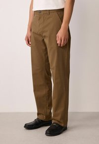 Bruine katoenen broek met een rechte snit, vlakke voorkant, twee zijzakken en een knoopsluiting. G gedragen met zwarte loafers.