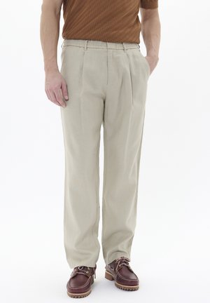 Pantalones beige con pliegues y cintura elástica. La tela parece ligera. Combinados con zapatos marrones con cordones y suela gruesa.
