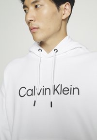 Witte hoodie met een groot zwart "Calvin Klein" logo. Inclusief trekkoorden met zwarte uiteinden en een gestructureerde stof.
