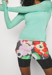 Hellblaues Langarmoberteil mit gemustertem Netzstruktur und mehrfarbigen Blumen-Shorts. Schwarze eng anliegende Shorts darunter. Grünes Akzentetikett.