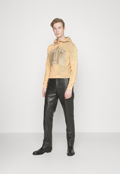Ανδρικό κίτρινο cropped φούτερ με υφασμάτινο σχέδιο και ασημένια γραφικά στοιχεία, συνδυασμένο με μαύρα δερμάτινα παντελόνια και μαύρα παπούτσια.