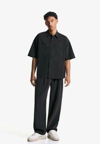 Black short-sleeve button-up shirt with two front pockets, paired with loose black trousers and white sneakers. Soft fabric with a relaxed fit.