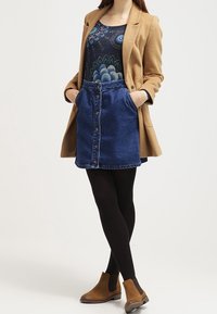 Beiger Blazer, gemustertes schwarzes Oberteil, Jeansrock mit Knöpfen vorne, schwarze Strumpfhose und braune Stiefeletten mit seitlichem Gummi.