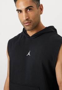 Sort ærmeløs hættetrøje med en frontlomme, der har et hvidt Jumpman-logo på brystet, lavet af blødt stof. Glat tekstur.