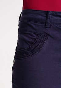 Marineblauwe broek met textuurdetails op de zakken, rechte snit en riemlussen. Gemaakt van zachte stof met een subtiele glans en stiksels.