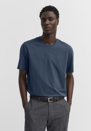 Junger Mann trägt ein blaues kurzärmeliges Hemd, das in graue Hosen mit einem geflochtenen dunklen Gürtel gesteckt ist, steht mit den Händen in den Taschen.