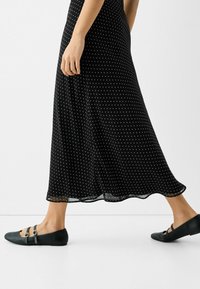 Czarna sukienka w białe kropki z zwiewnym dołem, połączona z czarnymi balerinami z dwoma klamrami. Gładka tekstura, minimalistyczny design i zaokrąglone noski.