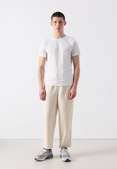 Jeune homme debout portant un t-shirt blanc uni, un pantalon large crème et des baskets grises et blanches sur un fond clair.