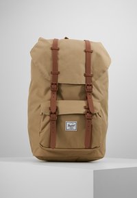 Beżowy plecak z płótna z brązowymi skórzanymi paskami, przednią kieszenią i górnym klapkiem. Posiada naszywkę z logo z przodu. Zbudowany z myślą o trwałości.