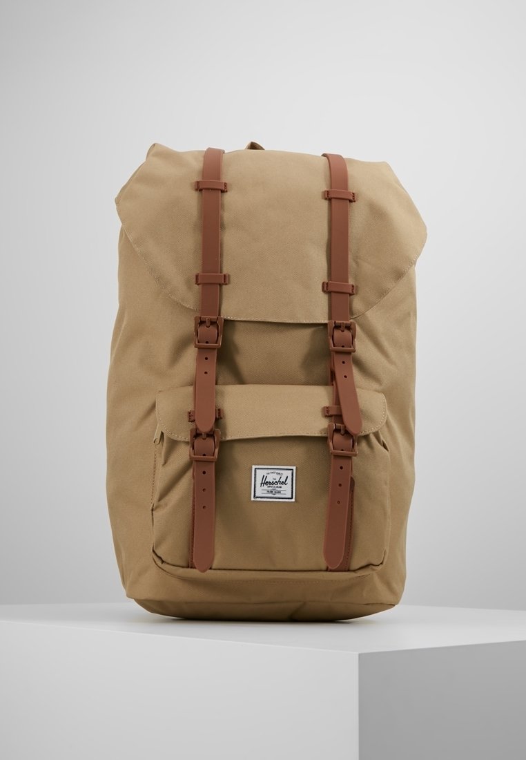 Beżowy plecak z płótna z brązowymi skórzanymi paskami, przednią kieszenią i górnym klapkiem. Posiada naszywkę z logo z przodu. Zbudowany z myślą o trwałości.