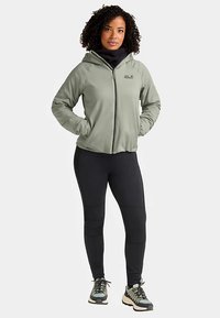 Groene jas met rits, gemaakt van gladde stof, gecombineerd met strakke zwarte broek. Opvallende kenmerken zijn een capuchon en twee voorzakken.