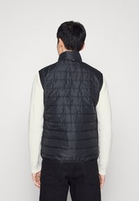 Sort puffervest med stående krave, der har horisontale stikninger og en glat, skinnende overflade, båret over en lysfarvet sweater.