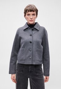 Casaco cinza curto com colarinho pontudo, fecho frontal de botões e dois bolsos grandes. O tecido parece texturizado com um acabamento suave.