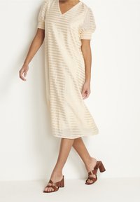 Cremefarbenes Midikleid mit V-Ausschnitt, kurzen Puffärmeln und vertikalen Streifen. Kombiniert mit braunen Absatzsandalen.