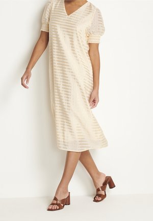 Robe midi couleur crème avec un col en V, des manches courtes bouffantes et des rayures verticales. Associée à des sandales à talons bruns.