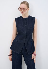 Chaleco de denim en azul oscuro con un dise�ño ajustado, sin mangas, con cierres de botones y bolsillos frontales, combinado con pantalones de pierna ancha a juego.