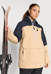 Bežová a námornícka lyžiarska bunda s prednou kapsou, zipsom a kapucňou na šnúrku, ktorú nosí osoba držiacou lyže. Má voľný strih.