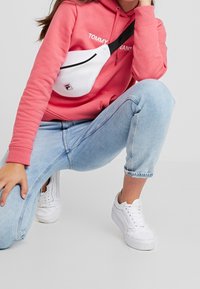 Bandolera blanca de malla con detalles en negro, llevaba sobre una sudadera rosa y jeans azules claros, con zapatillas blancas visibles.