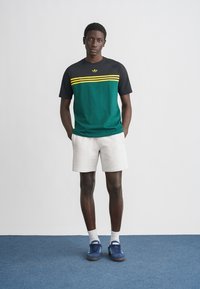 Žalia ir juoda trumpomis rankovėmis marškinėliai su geltonomis juostomis, derinami su baltais šortais ir mėlynais sportbačiais. Vienodas fonas, stovėjimo poza.