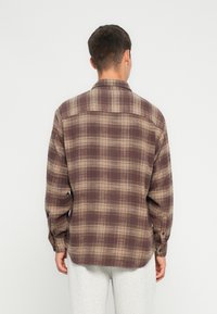 Brun rutete skjorte med lange ermer og krage med knapper. Har et strukturert stoff med lyse og mørke brune ruter og svake røde linjer.