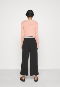 Rosa kabelstickad kort tröja, grå topp under, kombinerad med svarta vida byxor och svarta plattformsskor. Bakvy, neutral bakgrund.